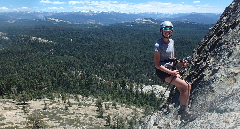 A young person wearing safety gear pauses to smile at the camera while rappelling a rock wall. There is a vast wooded and mountainous landscape in the background.